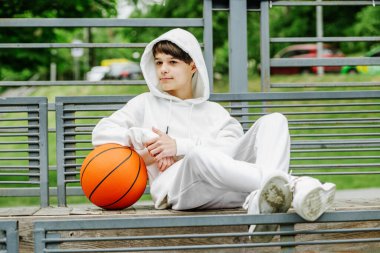 portrait of teenage boy sitting on bench with basketball in white tracksuit. Gen Z and sport concept.
