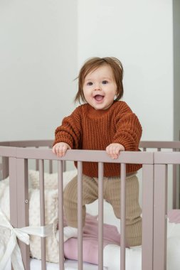 Cute laughing baby standing in round bed. Little girl learns to stand in her crib.