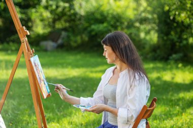 Young female artist paints picture in summer park in sunlight. Art and hobby concept. clipart