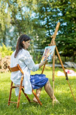 Young female artist paints picture in summer park in sunlight. Art and hobby concept. Back view