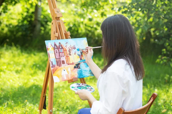 Young female artist paints picture in summer park in sunlight. Art and hobby concept. Back view