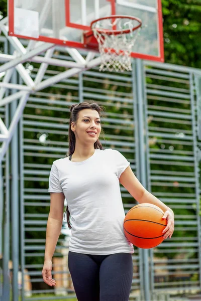 Concept of sports, hobbies and healthy lifestyle. Young athletic girl is training to play basketball on modern outdoor basketball court. Happy woman