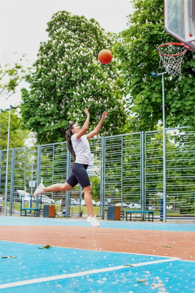 Concept of sports, hobbies and healthy lifestyle. Young athletic girl is training to play basketball on modern outdoor basketball court. Happy woman