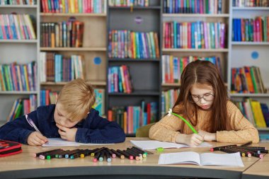 Schoolchildren write and draw sitting at table in library. Classmates study in classroom. Back to school