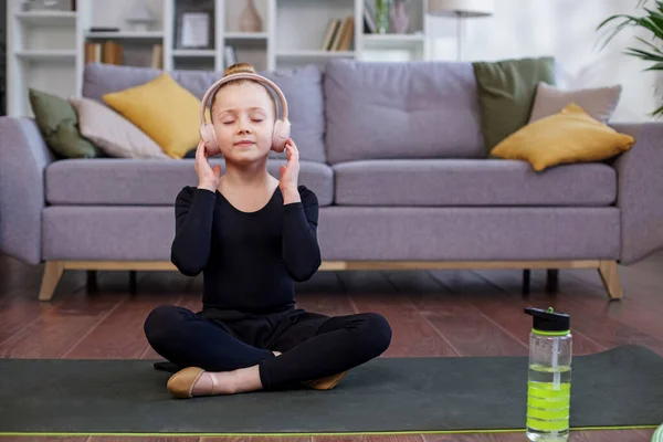 Child doing yoga exercise in room. Healthy lifestyle. Girl listens to music in headphones. Reusable water bottle.