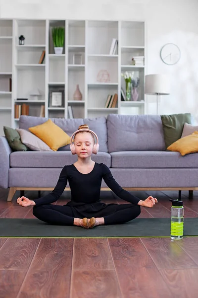 Child doing yoga exercise in room. Healthy lifestyle. Girl listens to music in headphones. Reusable water bottle.