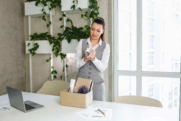 Dismissal. Frustrated fired caucasian employee woman packing belongings in cardboard box leaving workplace in modern office indoor.