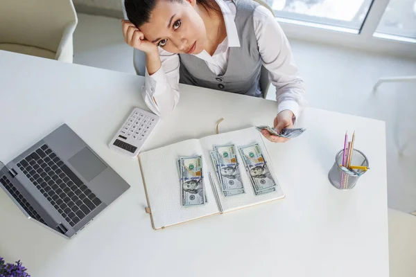 stock image Sad business woman sits at work desk in front of laptop and counts cash. Concept of low profits, low salary and financial problems. Top view.