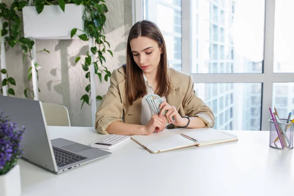 Sad business woman sits at work desk in front of laptop and counts cash. Concept of low profits, low salary and financial problems.