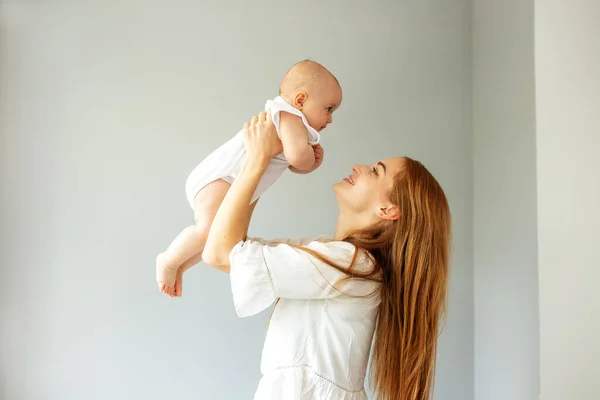 Happy cheerful young mother throws an adorable baby in air, plays, hugs baby with love, care, enjoys motherhood, maternity leave. Family spends time together.