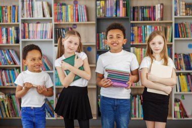Kütüphanede dört çocuk bir yığın kitap tutuyor. Çok ırklı sınıf arkadaşları eğleniyor. Dünya Kitap Günü. Günlük okumanın faydaları.