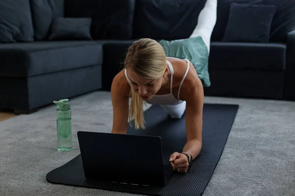 Training Concept. Fit woman doing yoga plank and watching online tutorials on laptop, training in room.