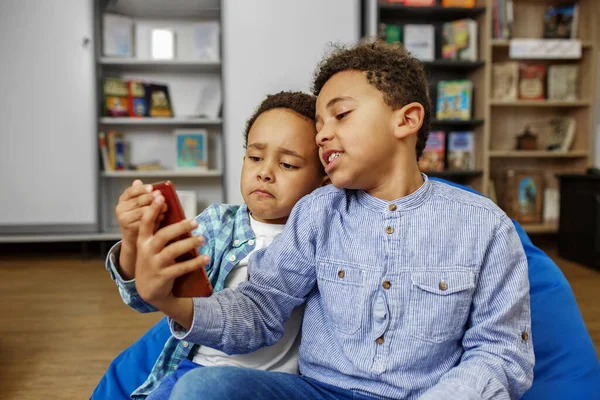 自宅でビーンバッグチェアに座っているときにスマートフォンで面白いビデオを見ている兄弟 ワイヤレス技術 — ストック写真