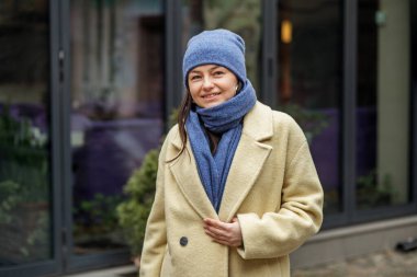 Şık, çekici, genç, güler yüzlü bir kadın kışın sokaklarda palto, mavi örgü şapka ve eşarp giyerek yürüyor. Mutlu bir ruh hali, moda modası.
