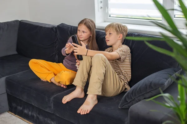 ソファーに座ってテレビの面白い映画を見ている子供たち テレビのリモコンを持っている少年 放課後に休憩する — ストック写真