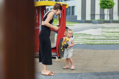 Anne, genç kızının oyuncak kamyonunu modern şehir parkında dikkatle izliyor..