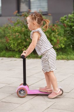 Çizgili elbiseli küçük kız pembeli scooterını dışarıda sürmeyi öğrenirken dikkatle odaklanıyor..