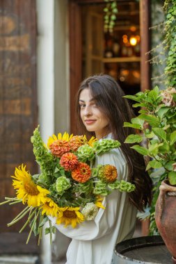 Yüzü dingin bir kadın elinde bir buket ayçiçeği ve canlı turuncu kasımpatı tutuyor. Şehir vahasında huzur dolu bir hayat sürüyor.