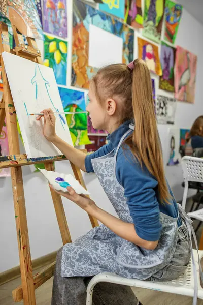 Focused young girl in an art class paints with brush on white canvas, surrounded by colorful artworks.