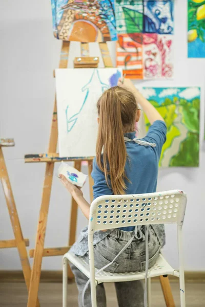 Focused young girl in an art class paints with brush on white canvas, surrounded by colorful artworks.