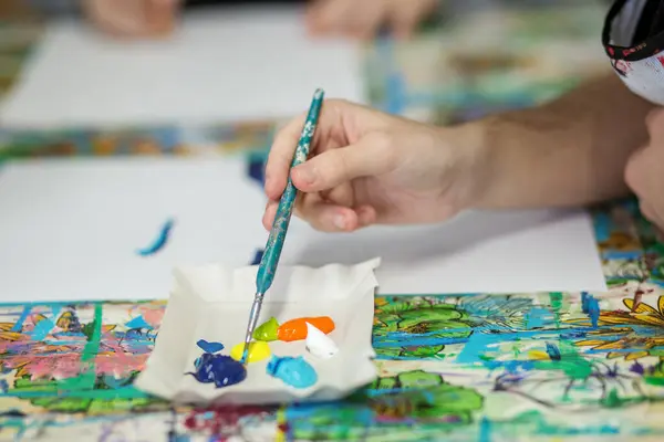 An artist\'s hand holding paintbrush over paper palette, selecting from vibrant blue, yellow, green, and orange acrylic paints for painting project.
