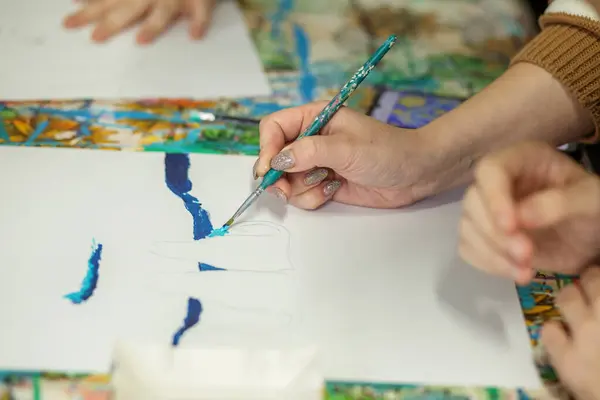 Close-up shot of an artist\'s hand applying strokes of blue acrylic paint to white canvas, showcasing creative process.