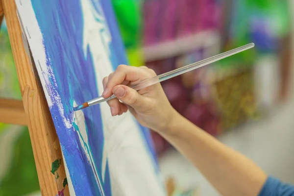 Focused artist\'s hand paints broad strokes of vivid blue on canvas, detailing motion and technique of painting
