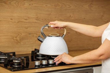 Kişi eli, sıcak mutfak atmosferinde sıcak içeceğe hazırlanmak için fırının üstüne şık beyaz çaydanlık koyuyor..