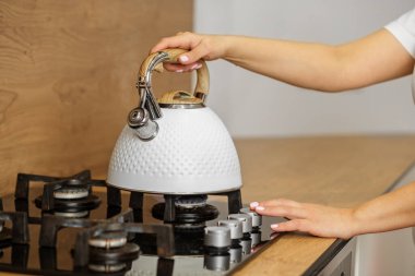 Kişi eli, sıcak mutfak atmosferinde sıcak içeceğe hazırlanmak için fırının üstüne şık beyaz çaydanlık koyuyor..