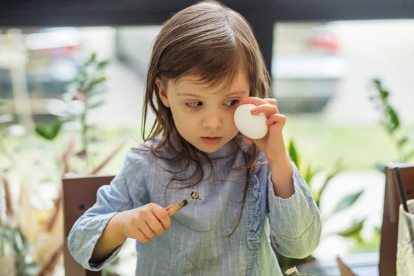 Focused child in blue dress paints an egg, traditional Easter activity.