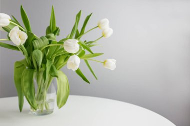 Beyaz bir masadaki cam vazoda duran bir buket beyaz lale minimalist ve huzurlu ev dekorasyonu manzarası sunuyor..