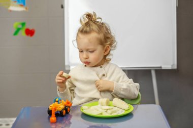 Anaokulu çocuğu renkli oyun masasında atıştırmalık molasında düşünceli bir şekilde muza bakar..