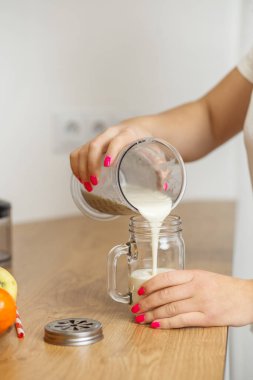 Elleri, mutfak tezgahındaki cam bardağa karıştırıcı kavanozdan kremalı bir smoothie dolduruyor. Arka planda meyve var..