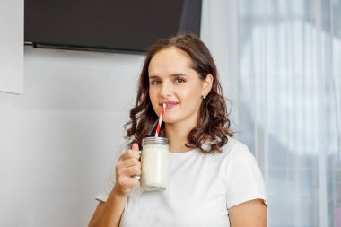 Beyaz tişörtlü memnun bir kadın modern mutfağında ev yapımı muzlu mascarpone smoothie yudumlamayı sever..