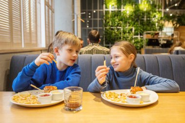 Mavi elbiseli, gülümseyen kardeşler restoran masasında oturup hamburger ve patates kızartmasının tadını çıkarıyorlar..
