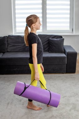 At kuyruklu genç bir kız evde mor yoga minderiyle yürüyor, spor yapmaya hazırlanıyor..
