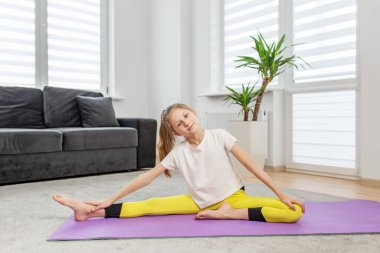 Öndeki küçük çocuk mor yoga minderine eğilip ev ortamında esnekliğe odaklanıyor..