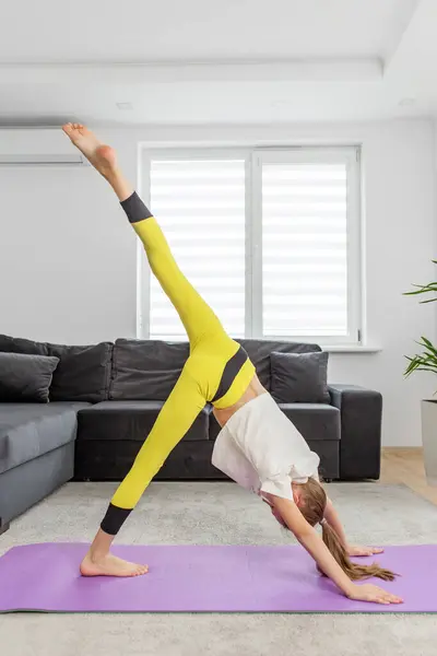 Child girl demonstrates an advanced yoga pose with one leg raised high, showcasing balance and flexibility in home environment.