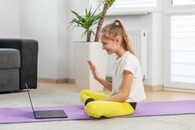 Yoga minderinde oturmuş gülümseyen reşit olmayan bir kız ev ortamında dizüstü bilgisayarındaki egzersiz dersini takip ediyor..
