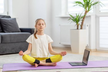 Serene çocuk nilüferde meditasyon yapıyor gözleri kapalı poz veriyor, parlak, huzurlu bir odada bilgisayarındaki bir derse katılıyor..