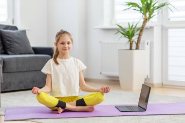 Serene kızı nilüfer çiçeği pozunda meditasyon yapıyor. Parlak, huzurlu bir odada dizüstü bilgisayarıyla bir derse katılıyor..