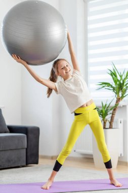 Neşeli kız yoga minderinin üzerinde durur, neşeli bir şekilde büyük bir spor topunu kafasının üzerine kaldırır, parlak, ev ortamında..