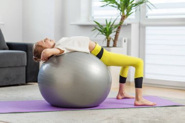 Genç bir kız spor salonunda arka eğimli oynuyor. Mor yoga minderiyle sırt kaslarını esnetirken rahatlıyor..