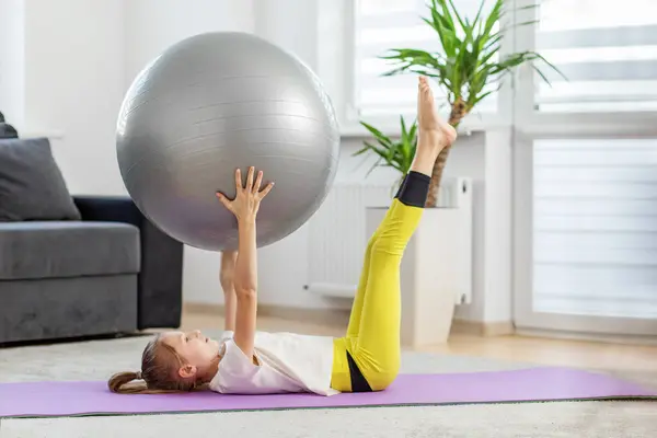 小女孩躺在家里的瑜伽垫上 用她的腿和胳膊提起一个健身球 进行核心强身健体运动 — 图库照片