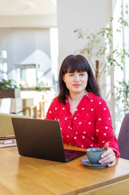 Sıcak bir gülümsemesi olan arkadaş canlısı bir kadın, samimi kafe ortamında, ahşap masada dizüstü bilgisayarıyla çalışıyor. Sıcak içecek..