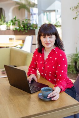 Sıcak tebessümü olan arkadaş canlısı bir kadın, rahat bir kafe ortamında, ahşap masada dizüstü bilgisayarıyla çalışıyor..