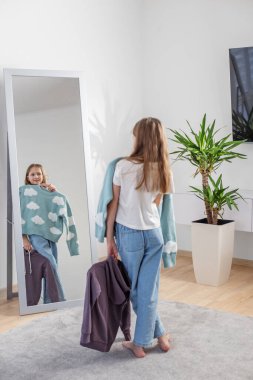 Kız giysi seçeneklerini inceler ve aynaya yansıtır. Etrafı ev bitkileriyle çevrili rahat bir oda ortamıyla çevrilidir..