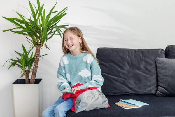 Joyful preteen girl laughs while holding tablet, seated next to red backpack on sofa in bright living room.