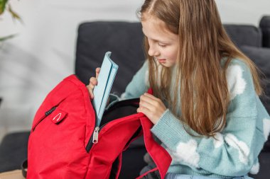 Kanepeye oturmuş, yanında bir kitapla kırmızı sırt çantasının içine bakıyor..