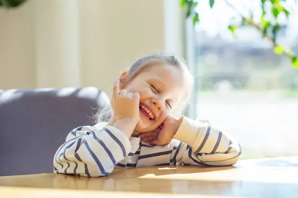 stock image Cheerful young girl laughing heartily while sitting at a wooden table bathed in sunlight.